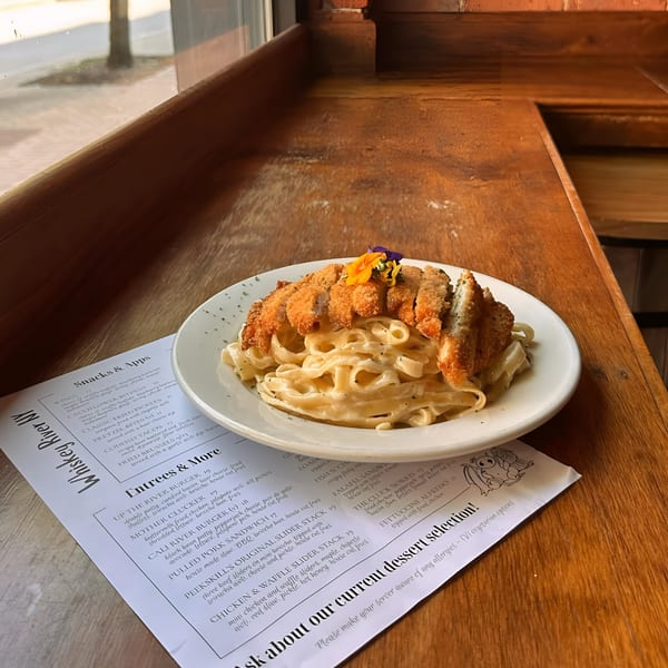 Whiskey River NY's Fettuccine Alfredo with fried chicken, garnished with edible flowers, on a wooden table.