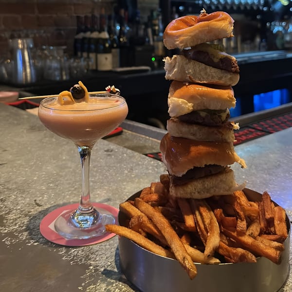 A tall stack of sliders and a side of fries next to a cocktail on a bar.